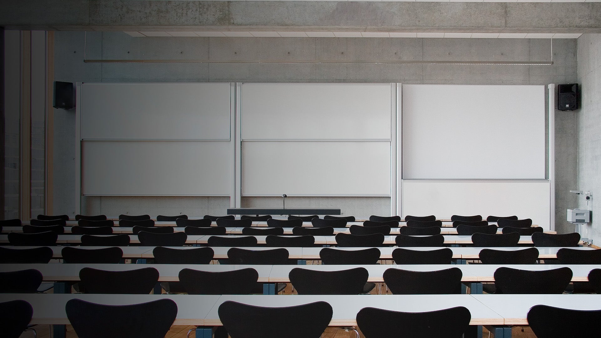 Spacious, well-lit modern classroom featuring rows of sleek black chairs and white tables, all oriented toward expansive whiteboards on a concrete wall—ideal learning space promoted by our expert classroom solutions company.