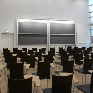 Modern classroom with black chairs, wooden desks, Chalk Column Board by Presentation Spaces, teacher's desk, and bright windows.