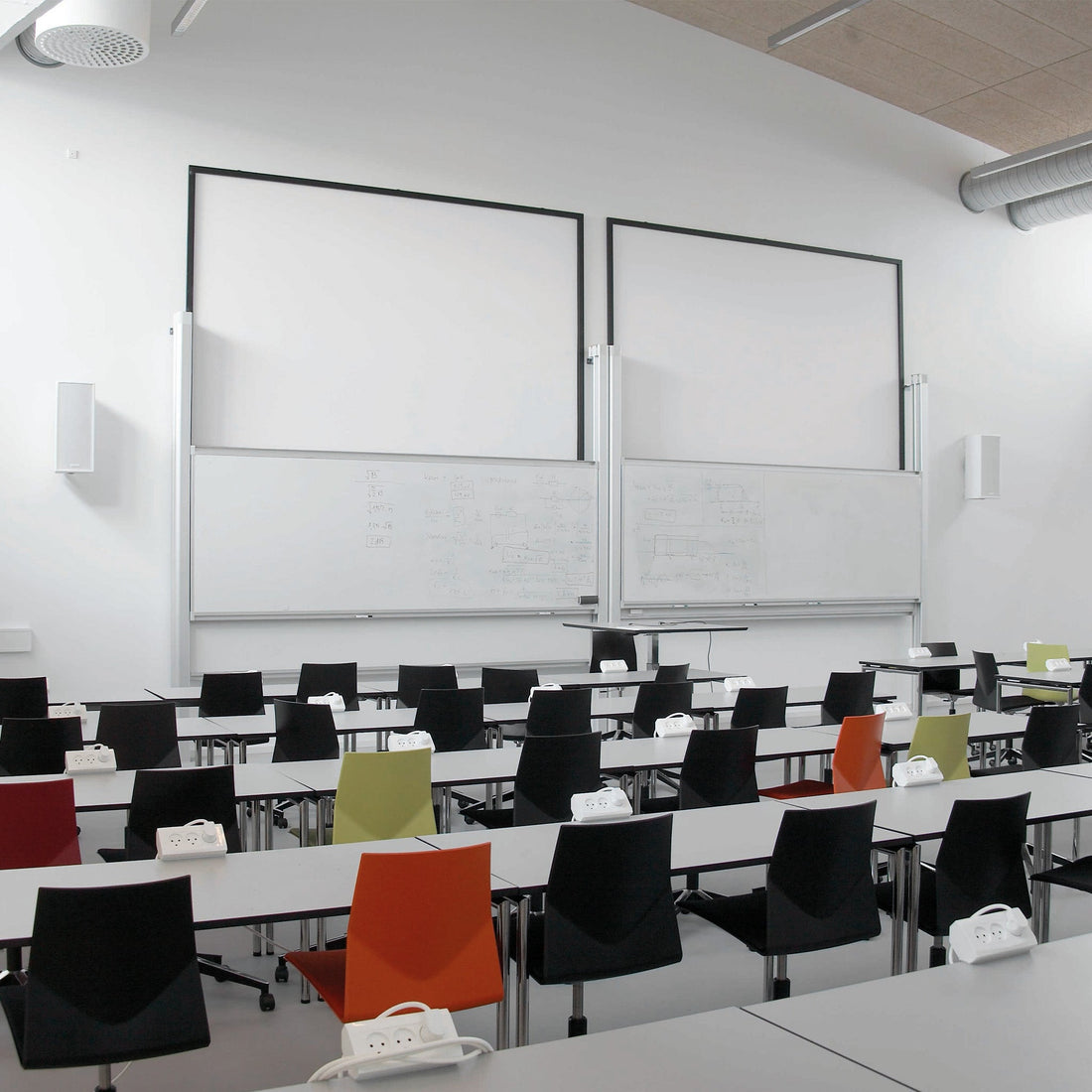 Spacious, well-lit modern classroom with colorful chairs, desks with outlets, and dual electric dry wipe boards with diagrams—empty room.