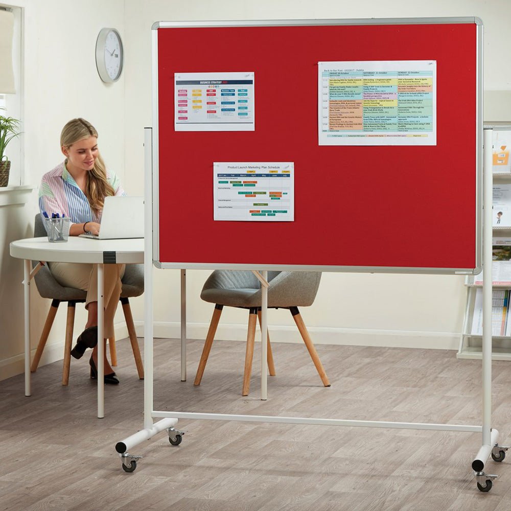 A professional woman works at a laptop near a height-adjustable double-sided felt notice board displaying charts and schedules.