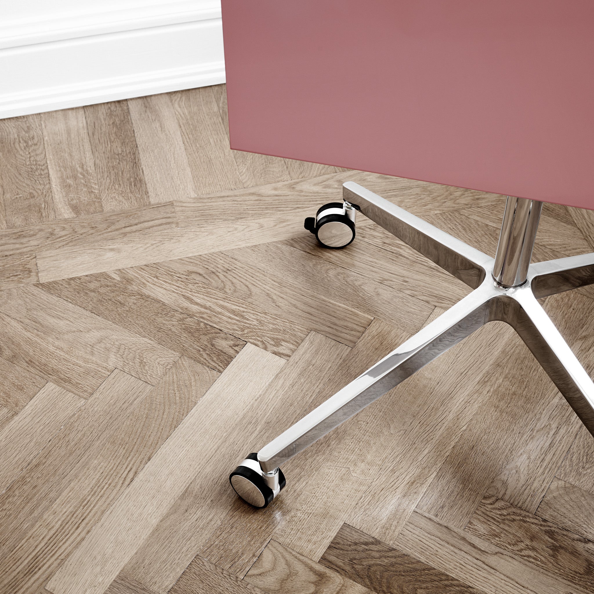 Close-up of Lintex Mood Mobile glassboard’s metal base and wheels, pink panel, on herringbone wood floor beside white wall.