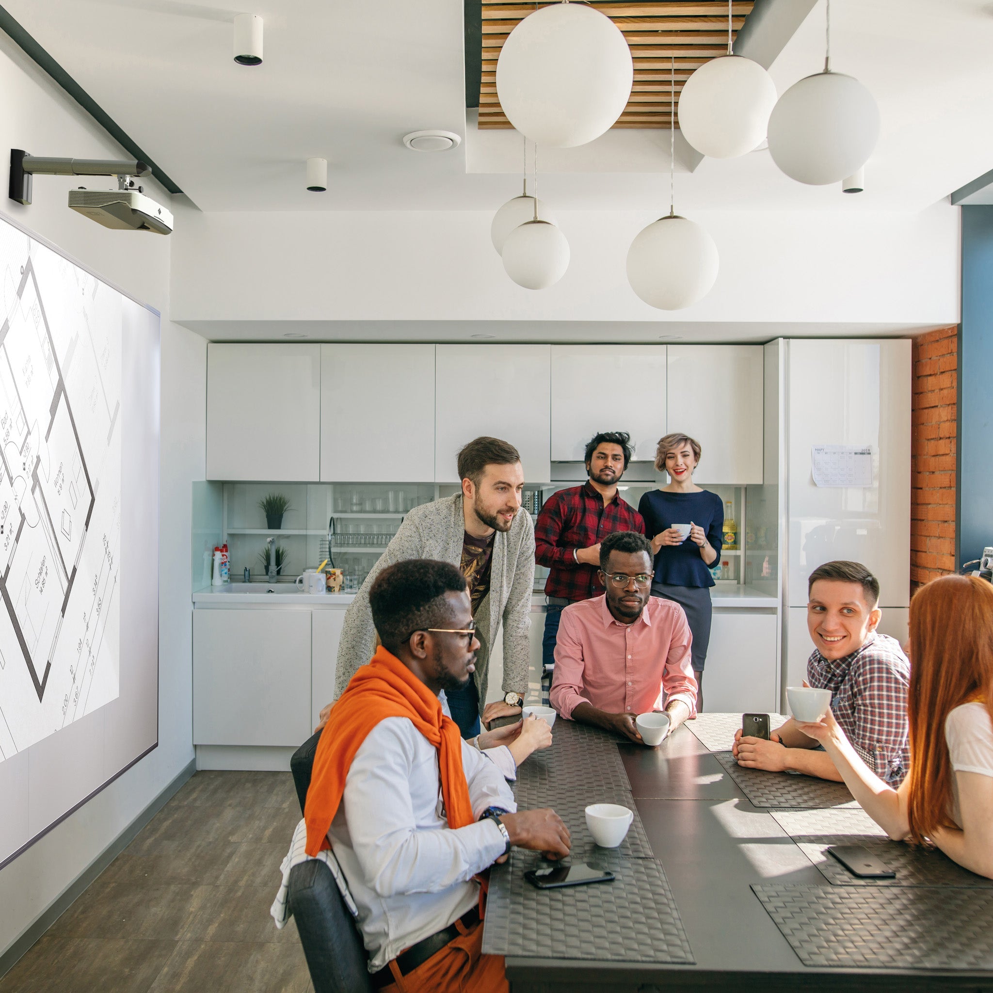 Six professionals discuss ideas in a sleek office kitchen, with laptops and coffee, near a Presentation Spaces Writing Wall display.