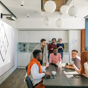 Modern office kitchen scene: six colleagues smiling, chatting by a table with coffee mugs; floor plan projected on writing wall.