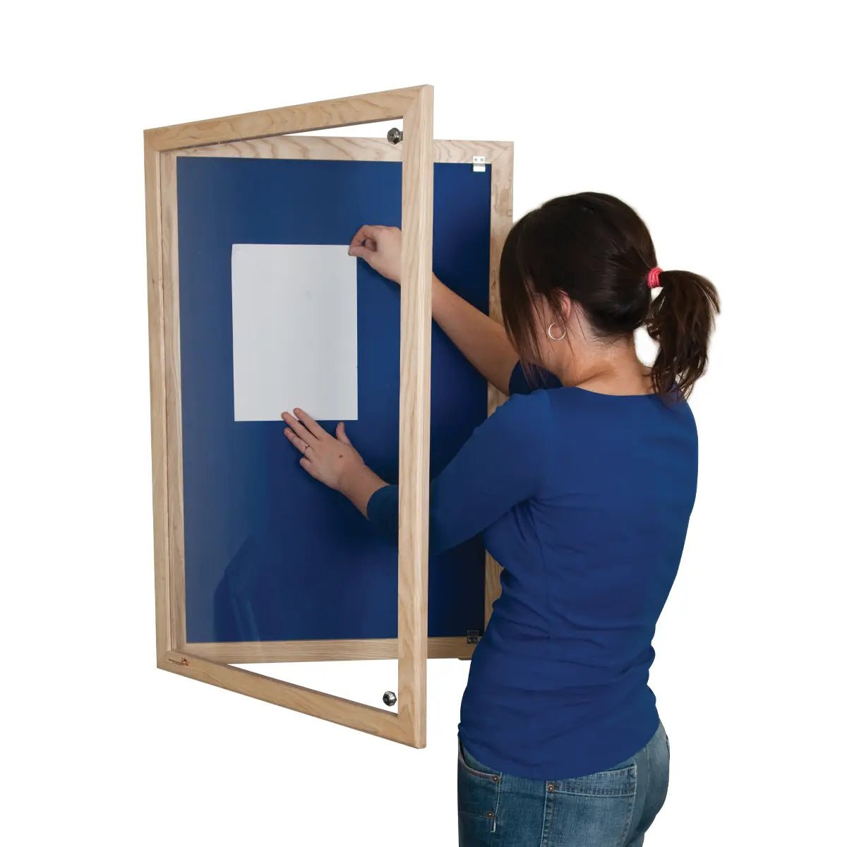 Woman places paper on Premier Felt Tamperproof Noticeboard by Presentation Spaces; ash frame, glass cover, blue interior shown.