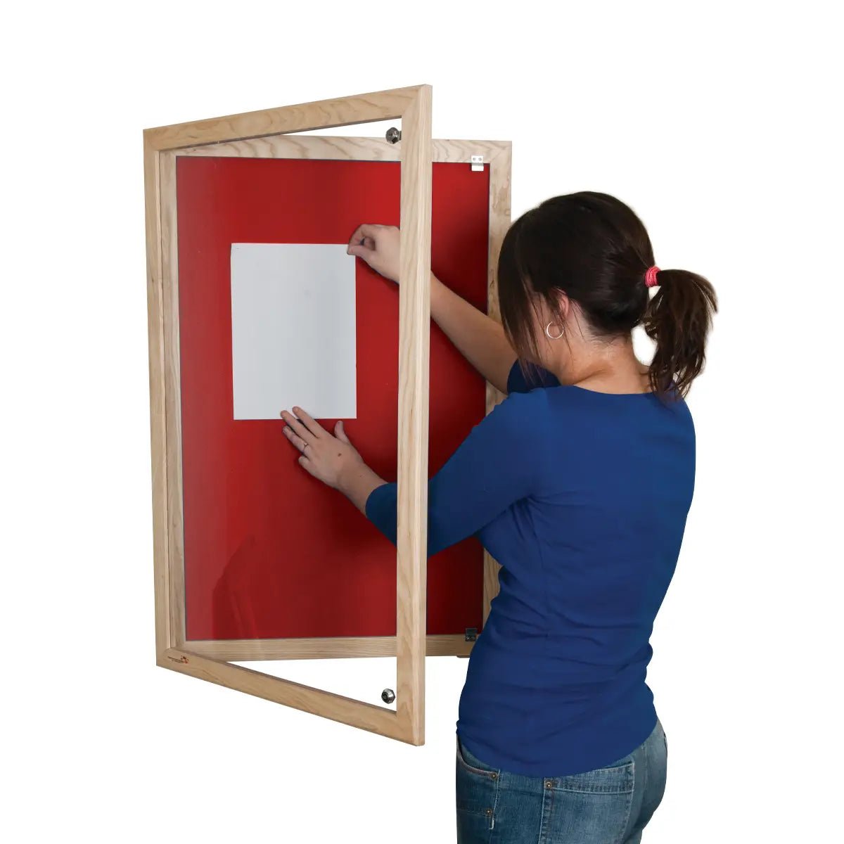 A woman pins paper to a Premier Felt Tamperproof Ash Hardwood Noticeboard, showcasing its red felt, glass door, and Class 1 Fire Rating.