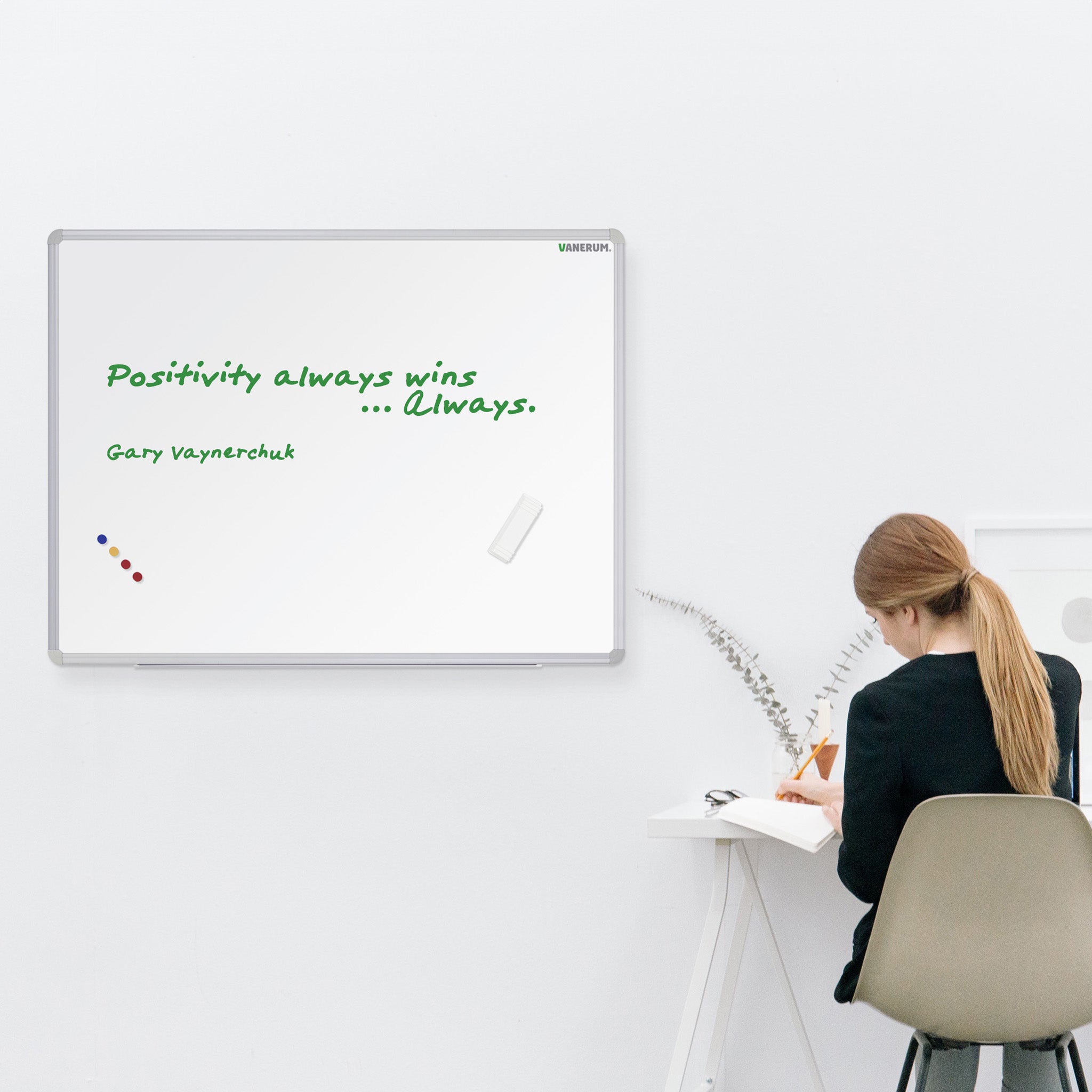 Woman with long hair writes at white desk; VES Magnetic Whiteboard displays “Positivity always wins... Always. Gary Vaynerchuk.”.
