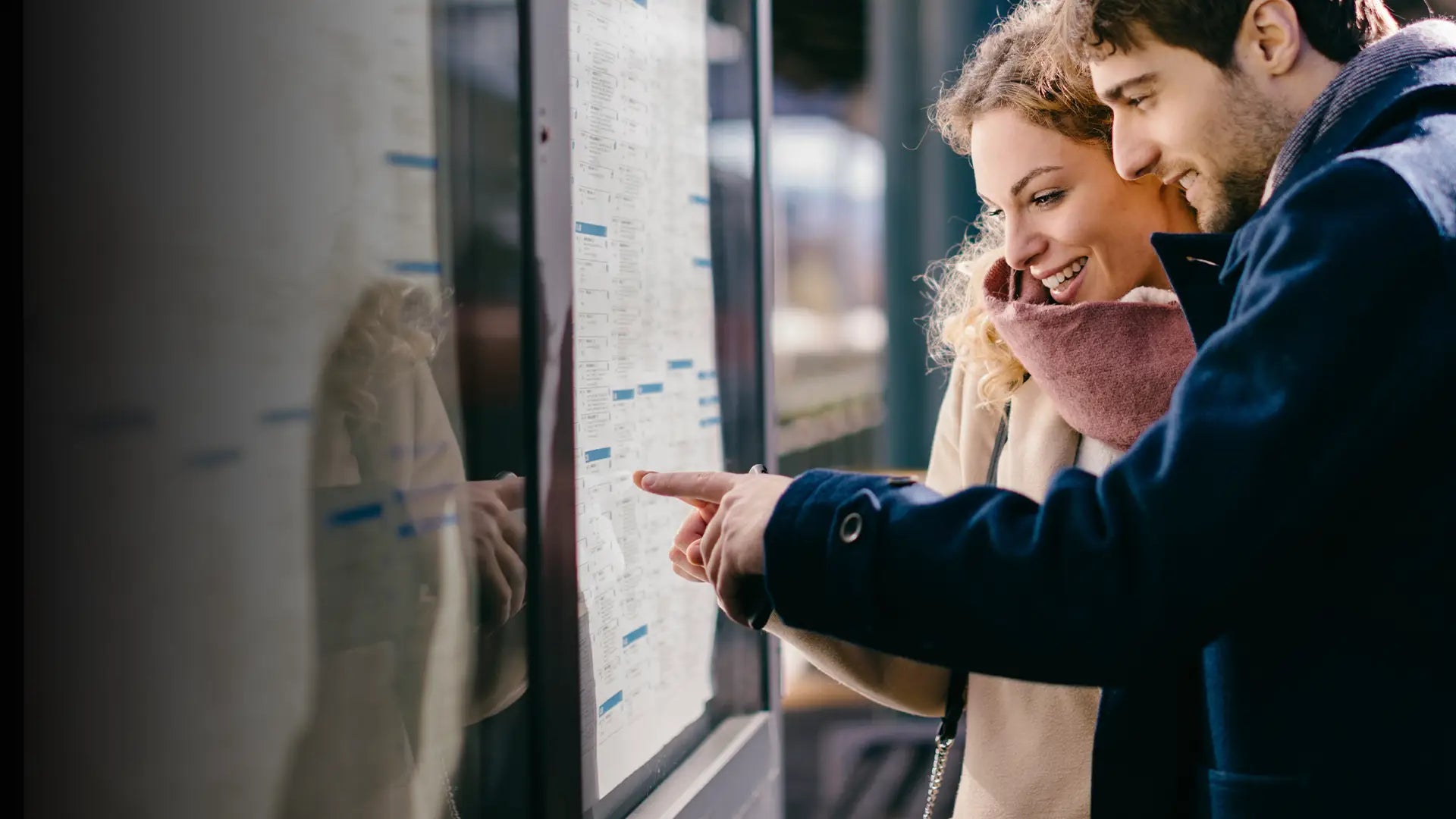 A happy couple in winter coats excitedly checks a public transport timetable on a glass display, engaging together as they plan their journey—perfect for trip planning and travel inspiration this season.